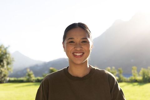 Sunlit asian woman smiling in park with mountain backlight wearing olive crewneck