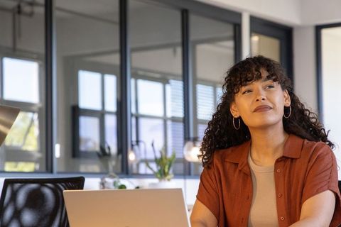 Thoughtful professional woman working on laptop in modern glass office with plants