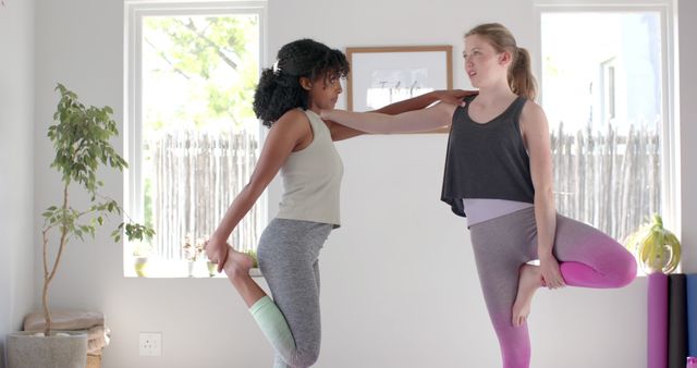 Two women practicing yoga together indoors in a bright room with natural light. One woman assists the other in holding a balancing pose. This image is ideal for depicting themes of fitness, wellness, and teamwork. Perfect for yoga studios, fitness blogs, and wellness websites emphasizing group activities and support in personal health journeys.
