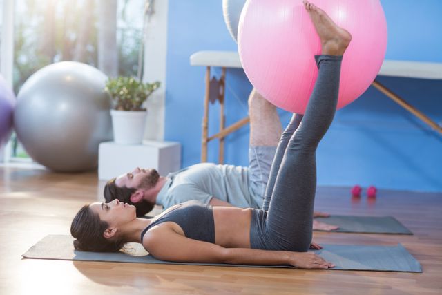 Man and woman lying on mats holding a stability ball between their legs in a clinic. This image can be used for promoting physical therapy, fitness routines, rehabilitation exercises, and healthy lifestyle choices. Ideal for websites, brochures, and articles related to health and wellness.
