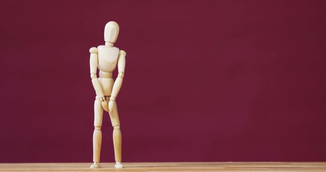 Wooden mannequin standing on a table against a red background with hands clasped in front. Useful for design projects, illustrating body language, art references, or educational materials related to human anatomy.