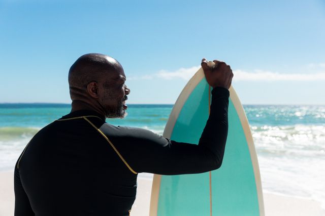 Rear view of african american senior man standing with blue surfboard at beach on sunny day. unaltered, active lifestyle, aquatic sport and holiday concept.