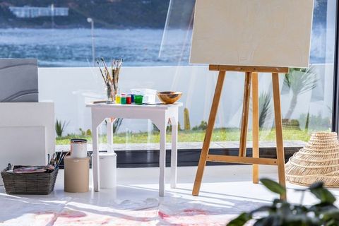 Sunlit coastal art studio featuring wooden easel holding primed canvas beside paint table