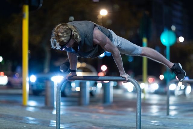 Front view of a fit Caucasian man with long blonde hair wearing sportswear exercising outdoors in the city at the evening, raising himself off the ground on a piece of street furniture, wearing head light.