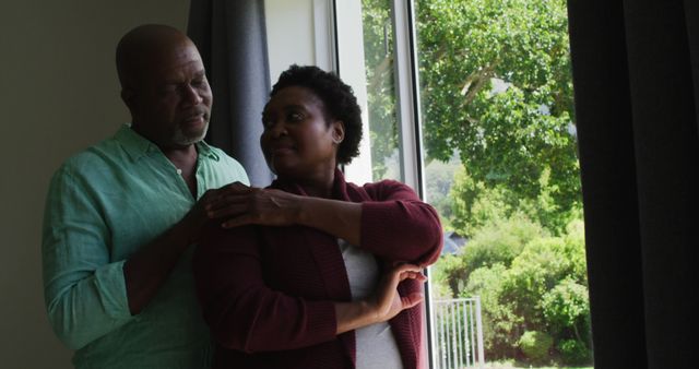 African american senior couple embracing each other while looking out of the window at home. retirement senior couple lifestyle living concept
