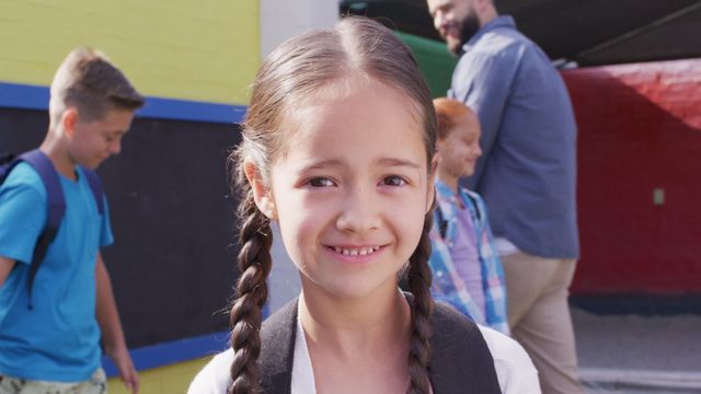 Smiling young girl with braided hair standing in the foreground with other children and male teacher in colorful school playground. Capturing the essence of back-to-school excitement and inclusivity. Ideal for educational content, back-to-school campaigns, and parenting or childcare articles.