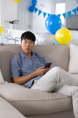Asian man sitting on sofa holding smartphone during casual home birthday celebration