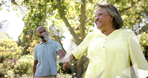 Senior couple walking hand in hand through sunlit park, joyful woman leading