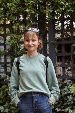 Smiling young woman standing in garden wearing green sweatshirt, backpack and sunglasses