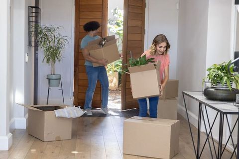 Couple carrying moving boxes and potted plants into bright sunlit foyer
