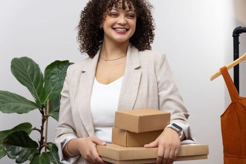 Smiling boutique owner holding stacked shipping boxes wearing blazer and smartwatch