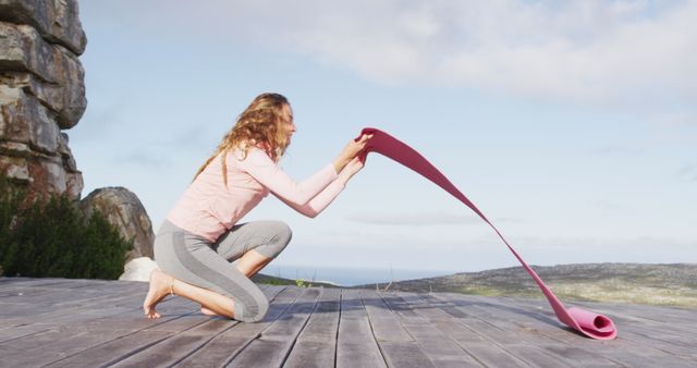Woman is unfurling a pink yoga mat on an outdoor deck with a scenic view, ready for a yoga or fitness session. This visual can be ideal for promoting wellness routines, outdoor activities, or yoga and fitness gear. The serene and natural setting emphasizes a healthy lifestyle.