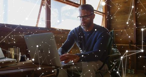 Mature man working remotely in rustic cabin typing on laptop with digital network overlay