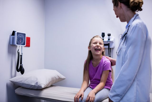 Doctor interacting with a smiling young patient in a hospital room. The child is sitting on an examination table, and the doctor is engaging with her, creating a comforting and friendly atmosphere. This image can be used for healthcare promotions, pediatric care advertisements, medical websites, and educational materials about doctor-patient relationships.