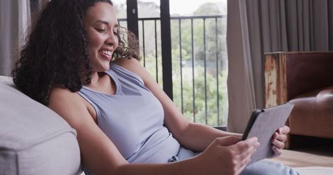 Smiling woman reclining on sofa scrolling tablet in sunlit cozy living room for relaxation