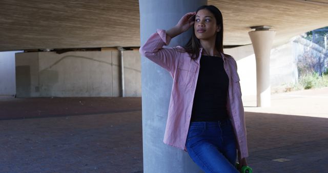 Young woman stands casually under an overpass, leaning against a concrete pillar. She wears jeans and a pink shirt, projecting confidence and contemplation. This urban scene is suitable for lifestyle blogs, fashion features, advertisements promoting youth apparel or urban exploration, and social media content.
