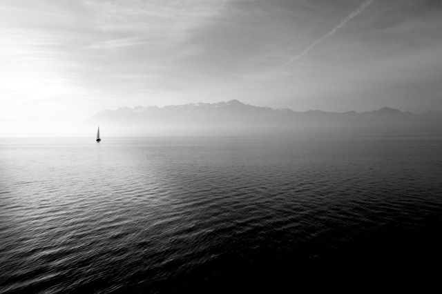 Silhouette of a small sailboat sailing on calm waters during sunrise with distant mountains visible through the mist. Excellent for use in articles or presentations about tranquility, solitude, nature, travel, or minimalist photography. Suitable for background images for websites, blogs, or brochures aiming to evoke peace and relaxation.