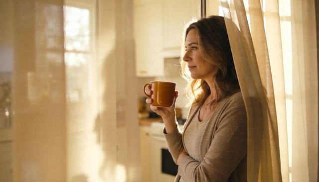 Middle-aged woman sipping coffee by sunlit window, cozy morning calm in home kitchen