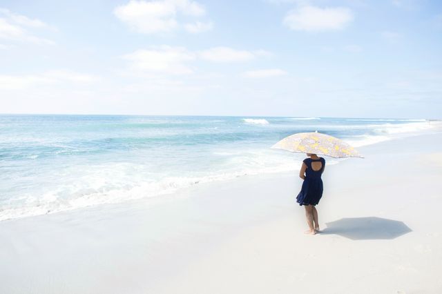 Woman stands on a quiet beach, shielding herself from the sun with an umbrella. Gentle waves roll onto the sandy shore. This can be used for vacation themes, peace and solitude promotions, or summer lifestyle blogs.