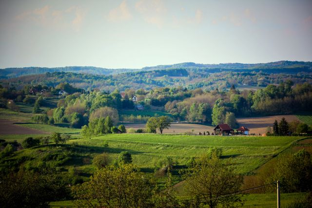Panoramic scene of lush green rolling hills bathed in natural light. Residential farmhouses and scattered trees dot the picturesque countryside, conveying a peaceful, rural atmosphere. Ideal for use in travel brochures, nature magazines, or promotional materials showcasing rural tourism and natural beauty.
