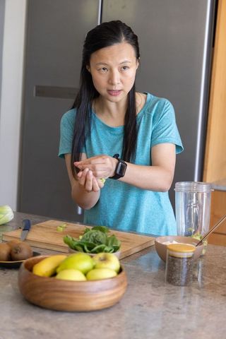 Asian woman preparing green smoothie in kitchen wearing smartwatch on wrist