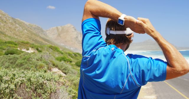 Man stretches on coastal road with ocean view, wearing blue athletic gear. Useful for promoting outdoor fitness, healthy living, exercise routines, athletic gear, adventure tourism, beach activities, and smartwatch technology.