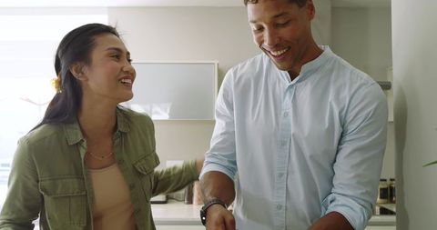 Sunlit diverse couple cooking and laughing in modern cozy kitchen, chopping vegetables