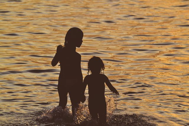 Children laughing and playing in shallow ocean waters during sunset. Golden hues of sky reflecting in water, creating warm evening light ambiance. Perfect for themes of childhood joy, beach vacation, family moments, summer activities.