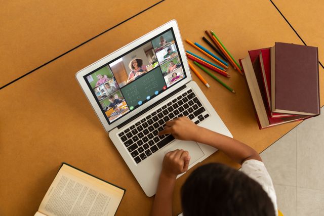 Child attending online class using laptop while sitting at table filled with books and pencils. Suitable for illustrating concepts of remote learning, virtual education, homeschooling, and modern educational practices.