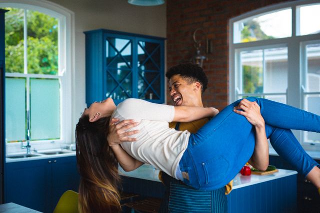 Young biracial man carrying his girlfriend in a modern kitchen, both smiling and enjoying their time together. Ideal for use in lifestyle blogs, relationship advice articles, home decor websites, and advertisements promoting happiness and togetherness.