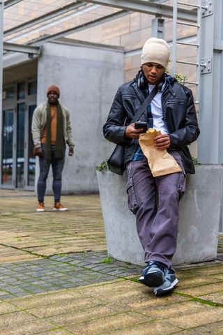 Young man using smartphone eating takeaway in urban courtyard with passerby walking