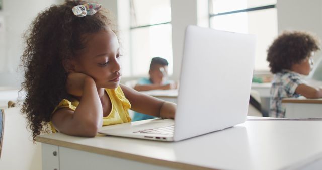 Image of bored african american girl sitting at desk with laptop during lesson in classroom. primary school education and learning concept.