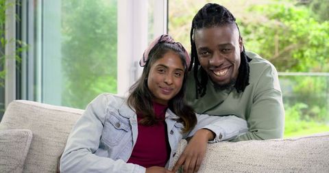 Smiling diverse couple sitting on sofa relaxing in sunlit modern living room