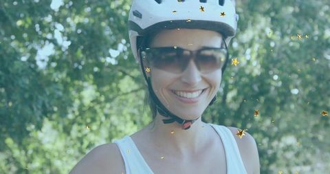 Smiling woman wearing bike helmet and sunglasses in sunlit park with gold star confetti