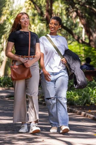 Two friends walking through sunlit park wearing casual outfits, glasses and crossbody bags