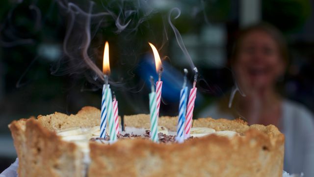 Colorful birthday cake with three lit candles partially blown out releasing smoke. Joyful event with blurred person celebrating in the background. Perfect for birthday greeting cards, party invitations, event promotions.