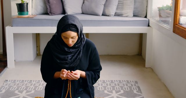 Muslim woman wearing a hijab praying with rosary beads in hands inside a modern, bright home with minimalistic furniture. This image is perfect for illustrating themes of spirituality, faith, and religious practices. Suitable for use in articles about Muslim culture, devotion, prayer routines, or interior design showcasing spiritual spaces.