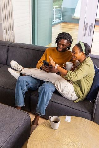 African american couple sharing smartphone and coffee on gray sofa at home