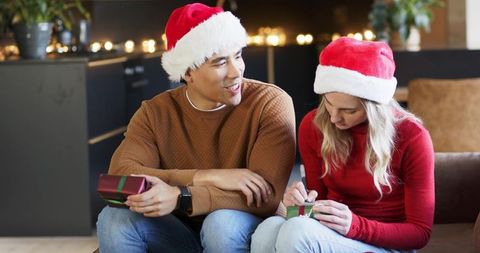 Couple wearing santa hats unwrapping gifts on cozy couch with warm holiday lights