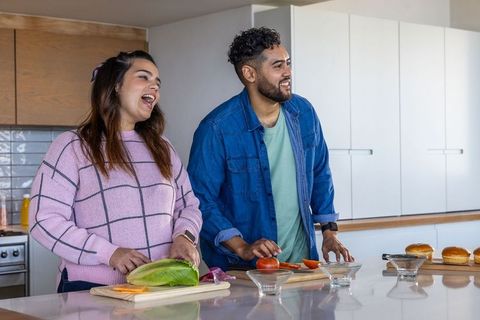 Laughing couple preparing fresh salad and burger toppings on marble kitchen island