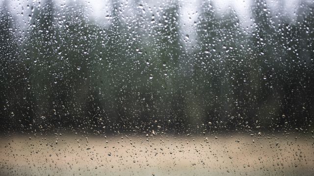 Raindrops adorning a window glass with a soft focus forest seen in the background. This image is ideal for conveying themes of nature, tranquility, introspection, or adverse weather conditions. Perfect for use in blogs, websites, weather reports, and inspirational projects.