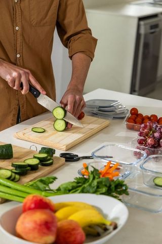 Slicing cucumber for meal prep on wooden board with fresh produce and storage containers