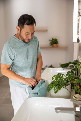 Mature asian man unzipping teal toiletry bag at bathroom counter preparing morning grooming