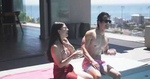 Asian couple laughing and sipping cocktails at rooftop pool on sunny sea-view terrace