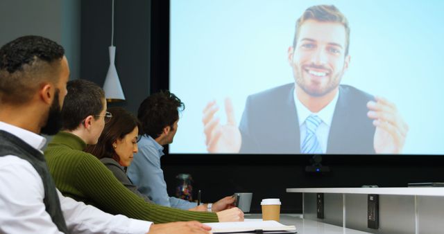 Team members watch a remote presentation on a video screen in an office conference room. Perfect for topics on remote work, business meetings, corporate presentations, and teamwork dynamics.