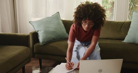 African American woman working from home writing notes while using laptop on sofa