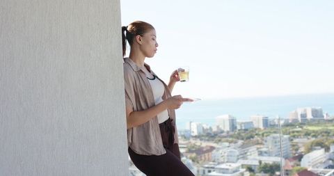 Leaning on balcony railing holding glass cup and saucer overlooking coastal city skyline