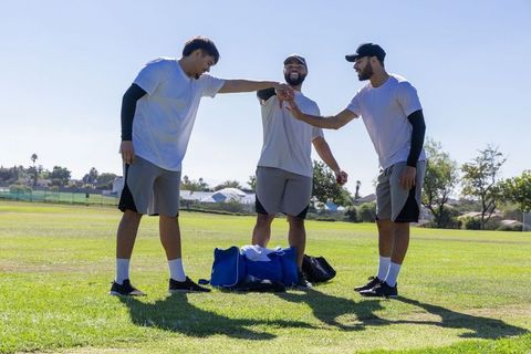 Male teammates joining hands in pregame huddle on sunny grass field with duffel bag