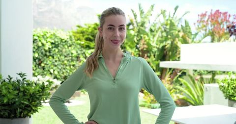 Young woman standing on patio wearing light green pullover smiling among backyard greenery