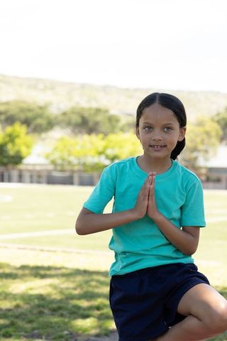 School-age girl practicing tree pose balancing on one leg on sunny school sports field